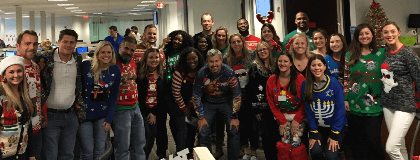 photo of several Allegis Group employees wearing holiday sweaters and costumes smiling together for a group picture in an office